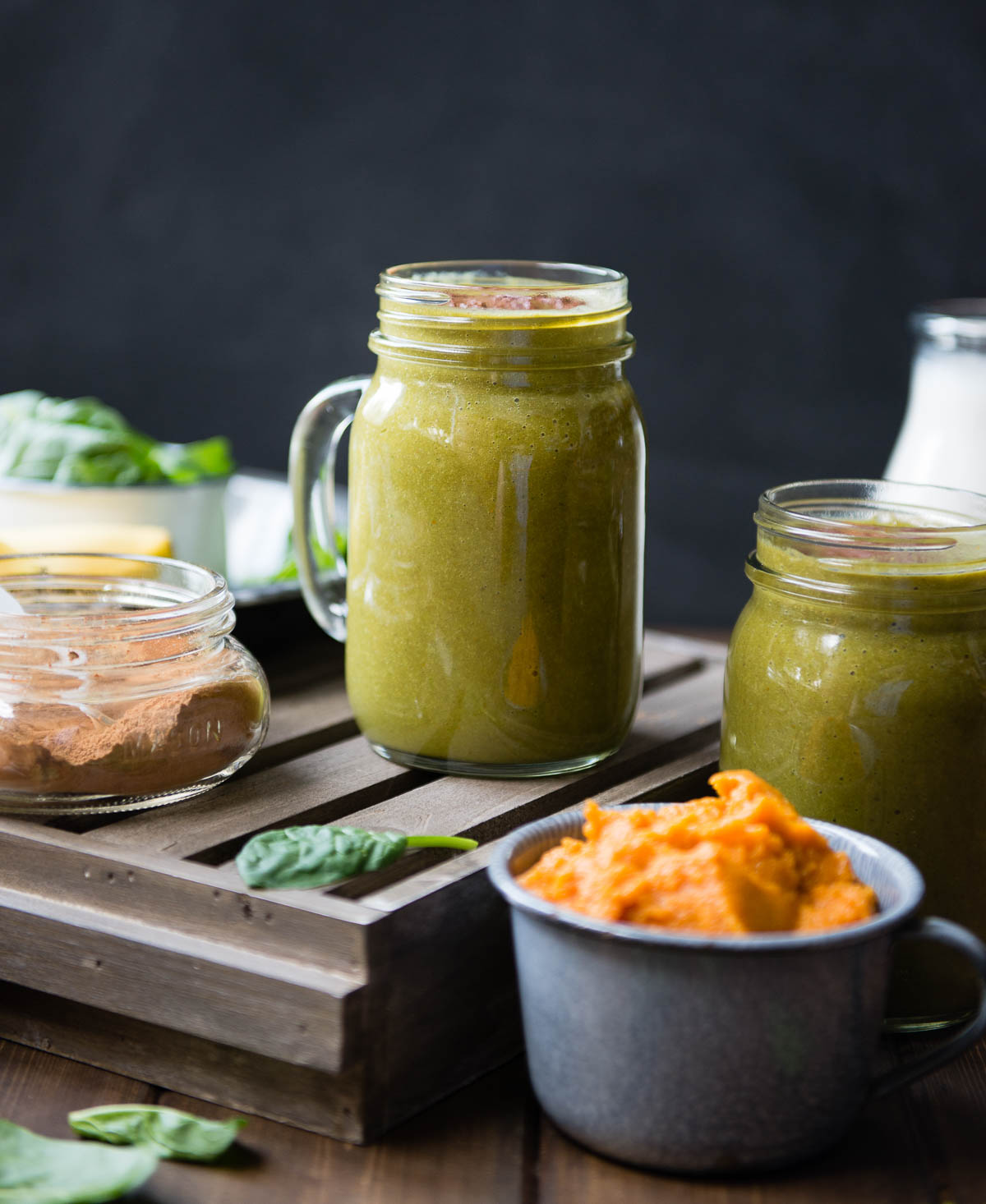 glass mug of pumpkin pie smoothie on a wooden tray next to a jar of spice and a tin cup of pumpkin puree.