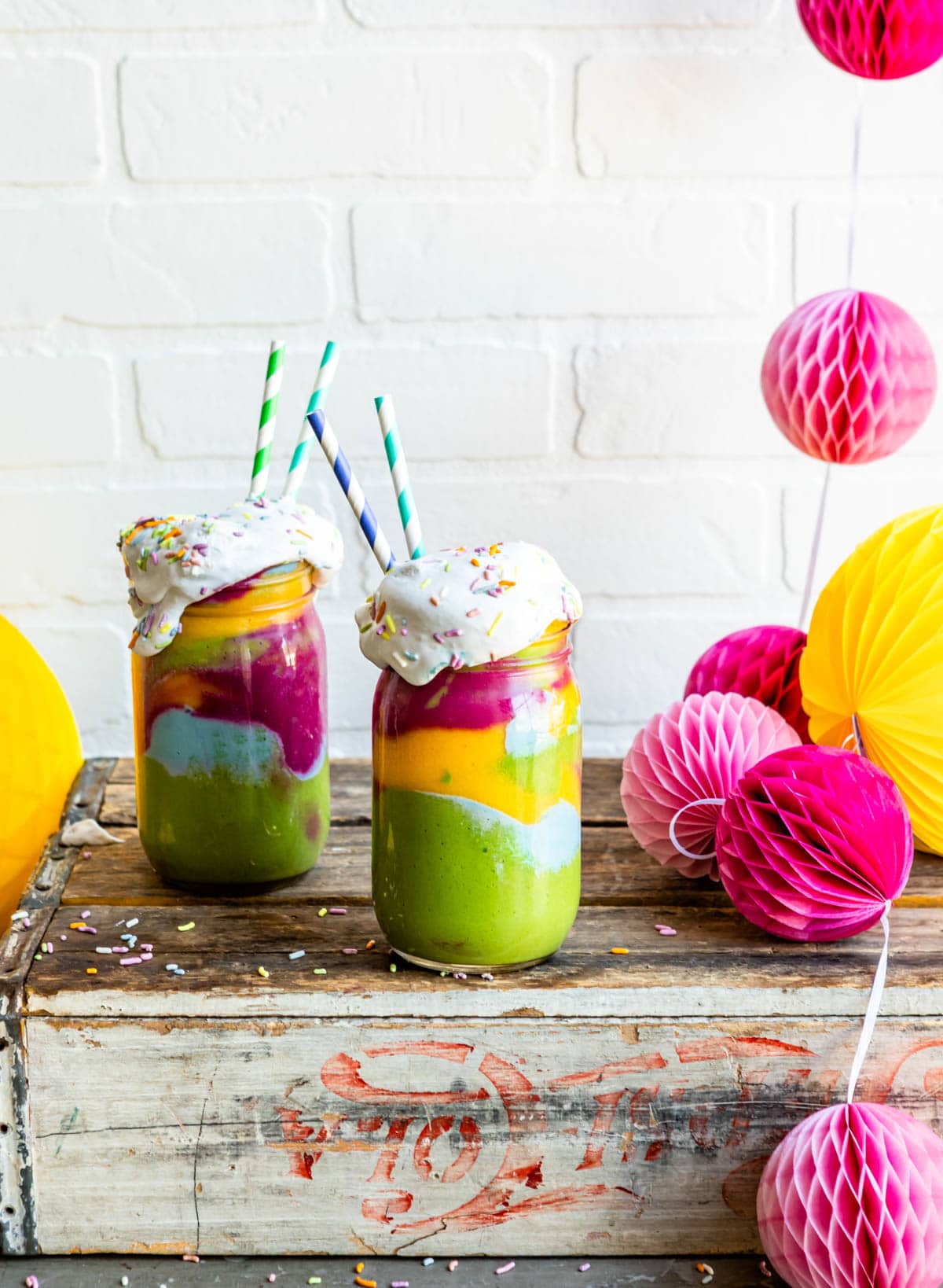 rainbow smoothie to share on a counter with party decorations and whipped cream + sprinkles to top.
