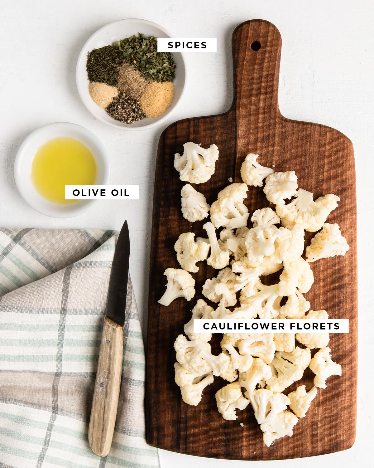 labeled ingredients for healthy snack including spices in a white bowl, olive oil in similar bowl and cauliflower florets on a wooden cutting board.