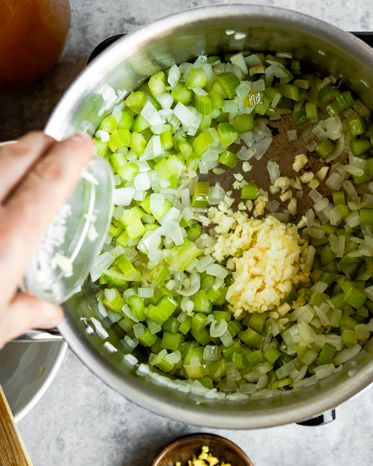 a persons hand adding garlic and ginger to sautéing onion and celery
