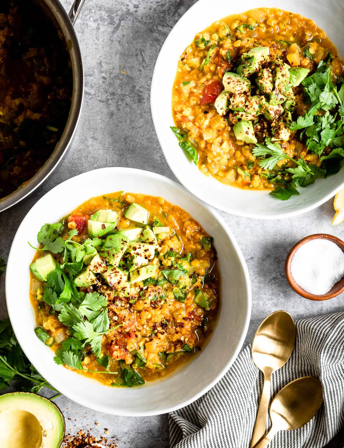 2 white bowls of red lentil soup ready to be eaten topped with fresh parsley, next to a small bowl of salt and a golden spoon.