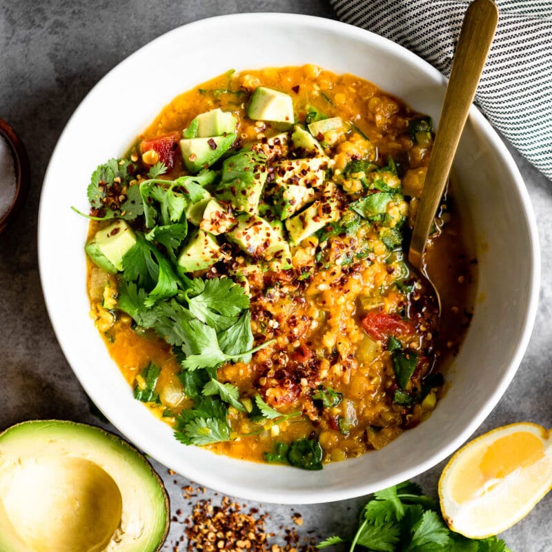 red lentil soup in a white bowl, topped with fresh avocado and a gold spoon.