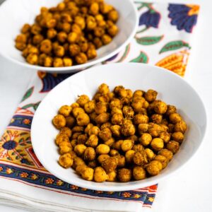 2 white bowls of roasted chickpeas snack on a colorful tea towel.