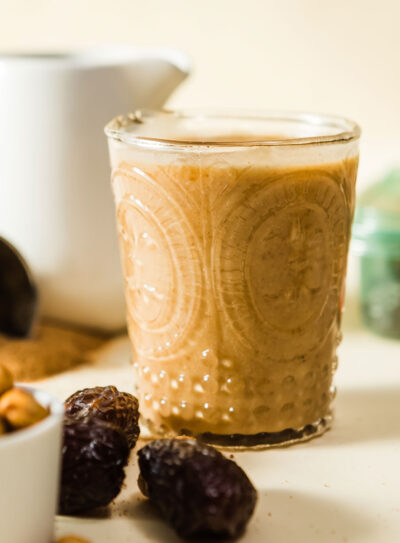 Salted caramel protein shake in a festive etched glass next to a bowl of cashews and some pitted medjool dates.