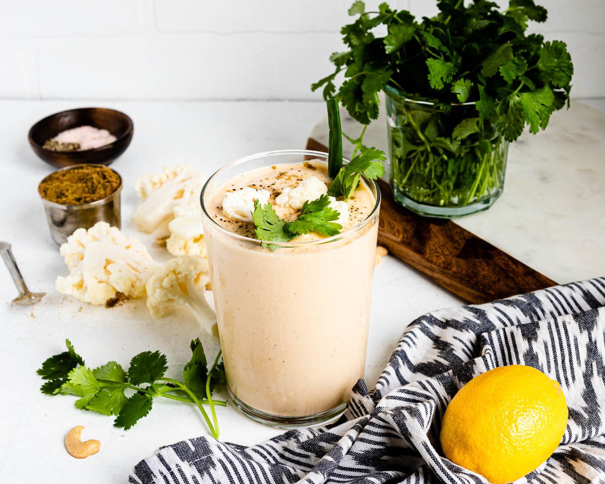 smoothie with cauliflower in a large glass topped with seasoned cauliflower florets, green onion and herbs on a white counter top.