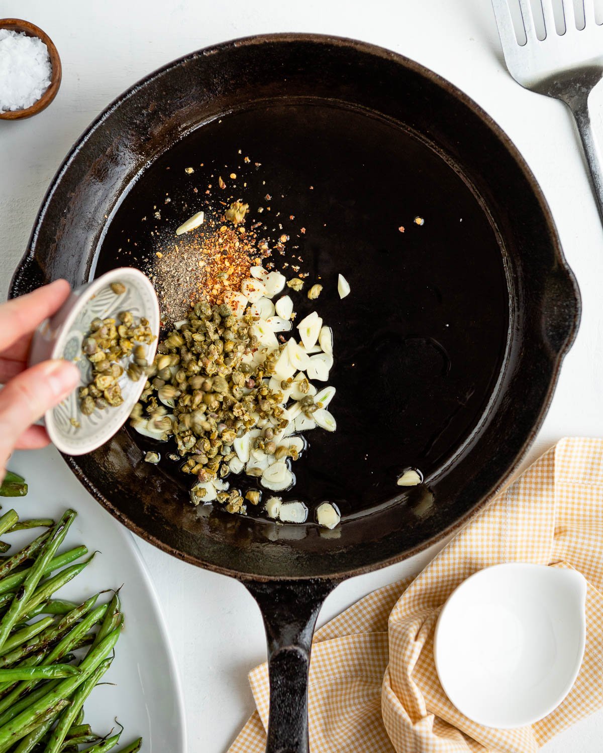 Adding capers, garlic, and spices to a cast iron skillet.
