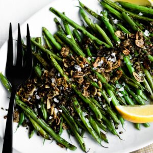 A close-up image detailing a black serving fork, skillet green beans with fresh sautéed topping, and a lemon wedge for garnish.