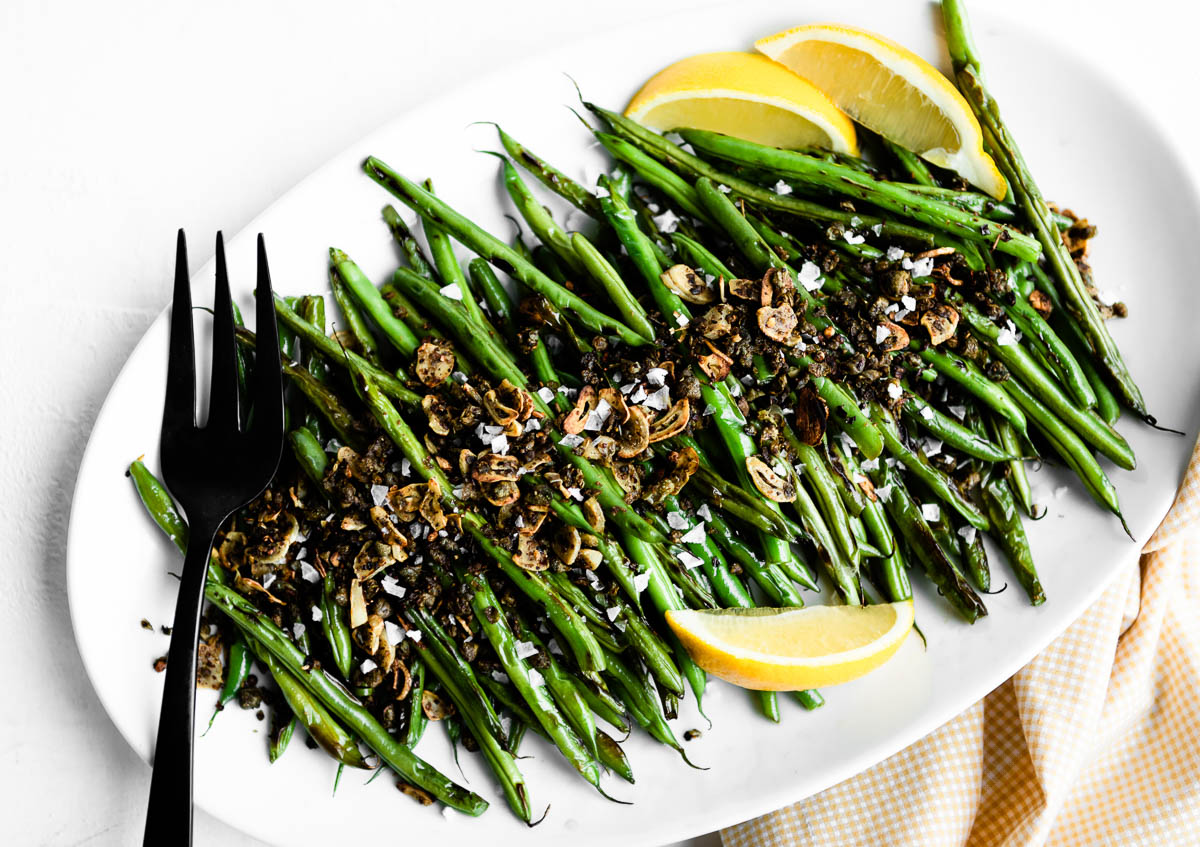 sautéed green beans on a white serving tray with a black fork, topped with capers and garlic, with lemon wedges on the side.