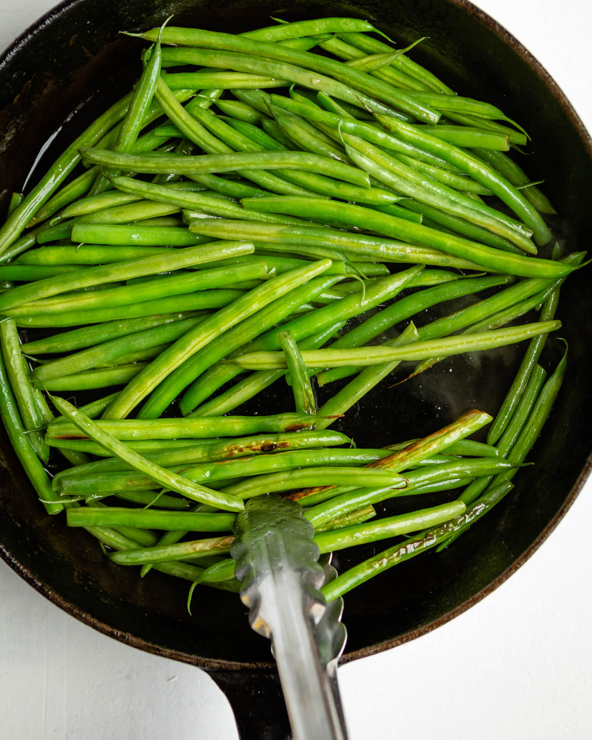 sautéed green beans in a cast iron skillet with metal tongs.