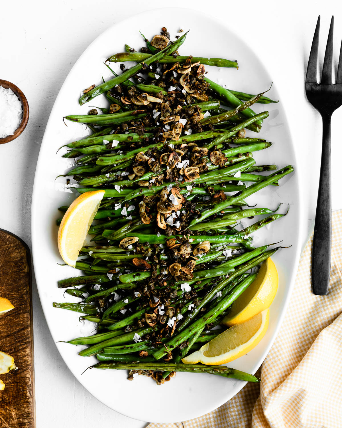 green beans topped with capers, garlic and spices on a white serving tray with lemon wedges.