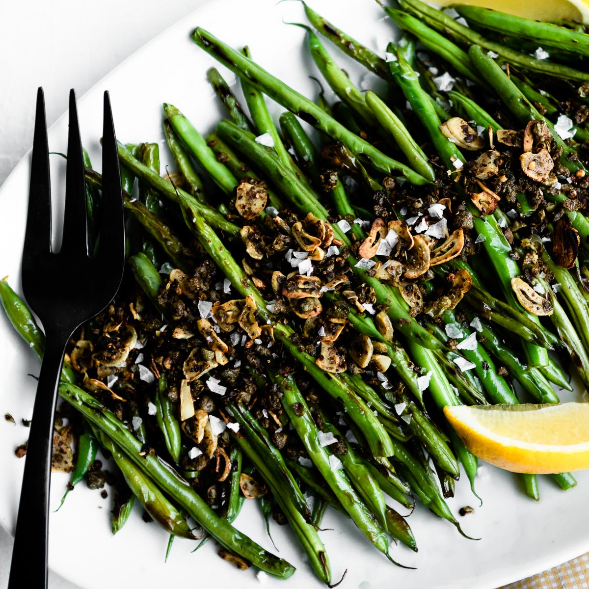 blackened green beans on a white serving tray topped with capers and garlic.