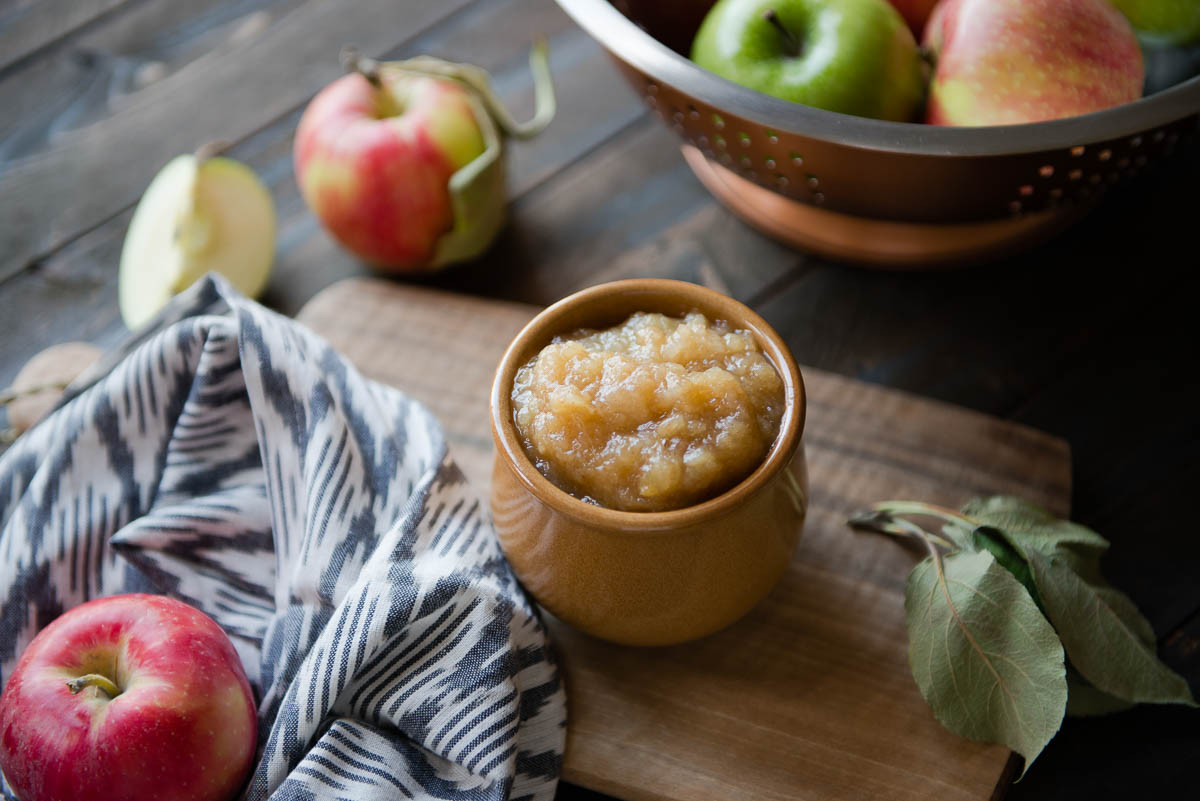 homemade applesauce in a brown ceramic bowl next to a patterned tea towel on a wooden cutting board.