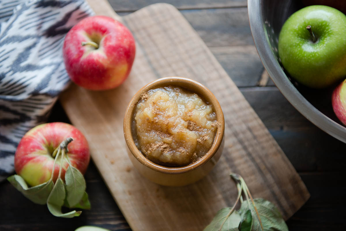 brown bowl of cooked apples on a wooden cutting board surrounded by fresh red and green apples.