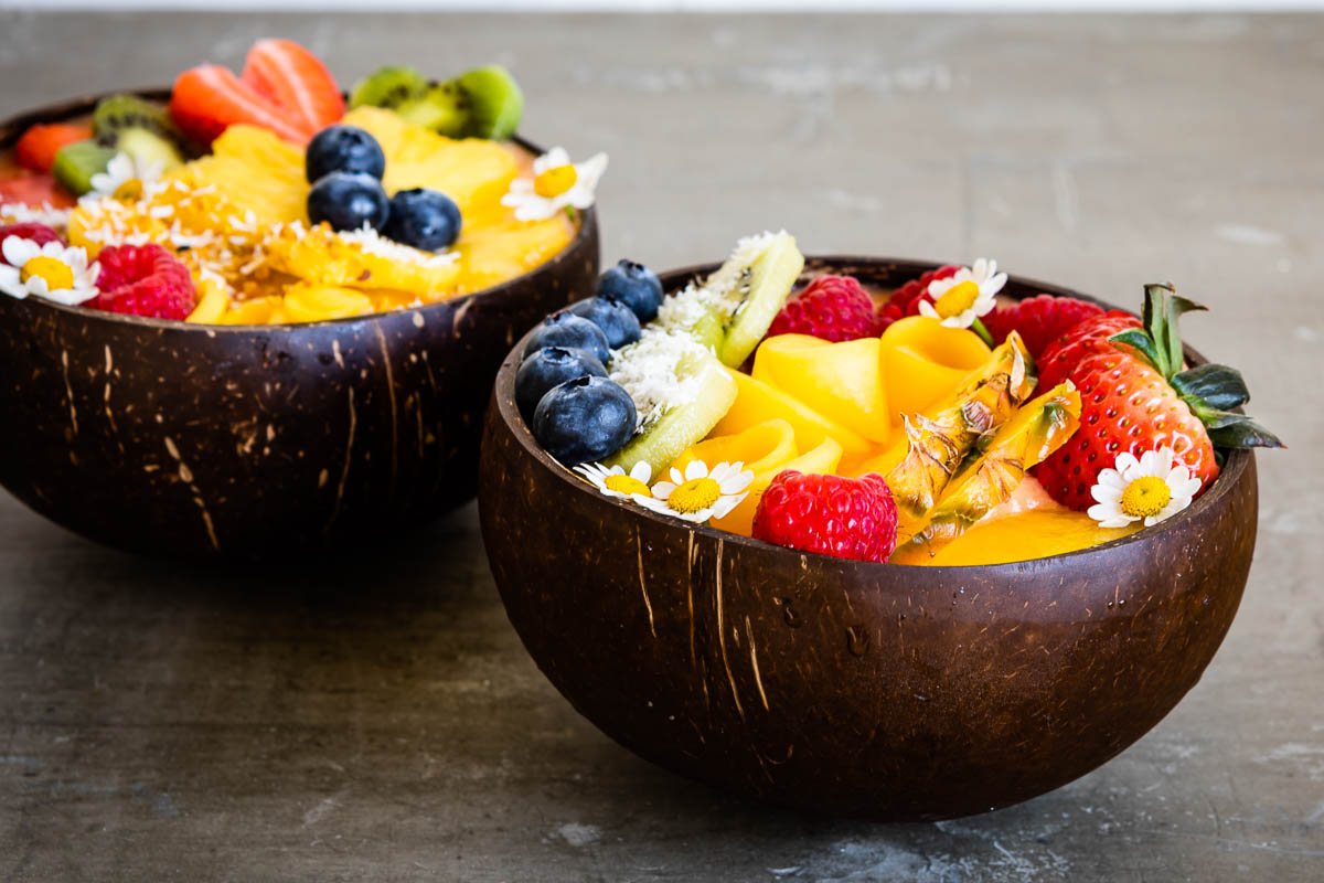 2 coconut bowls showing smoothie bowl recipes topped with a variety of fresh fruit and flowers.