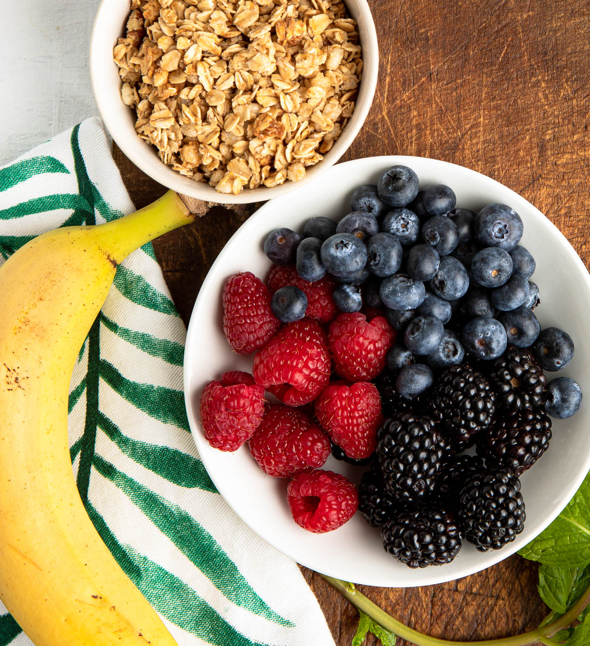 2 white bowls, one with granola and one with a variety of fresh berries, next to a whole banana.