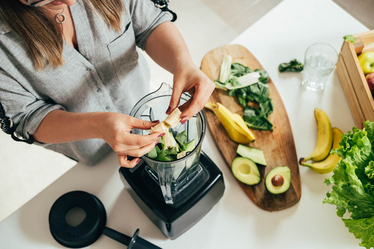 2 hands breaking banana over a blender container next to a wooden cutting board of fruits and vegetables.