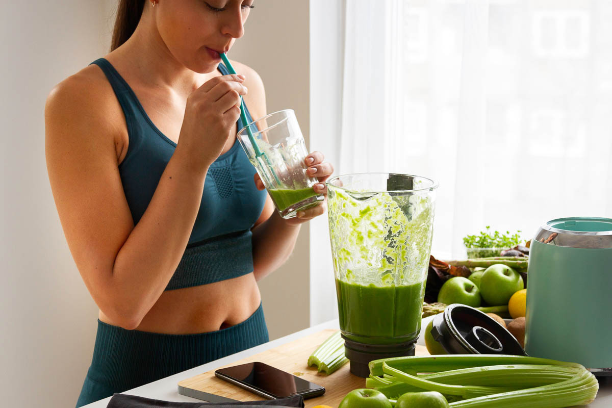 person in workout clothes sipping a green smoothie next to a blender of green smoothie in a kitchen surrounded by green produce.