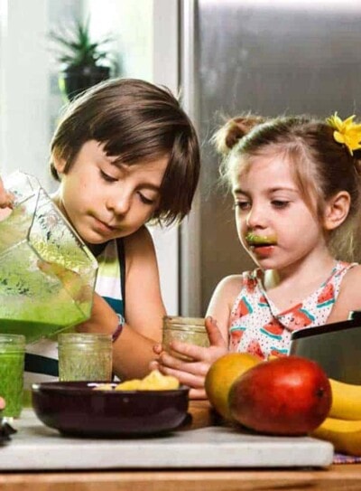 two kids pouring a green smoothie for kids surrounded by a counter full of fresh produce