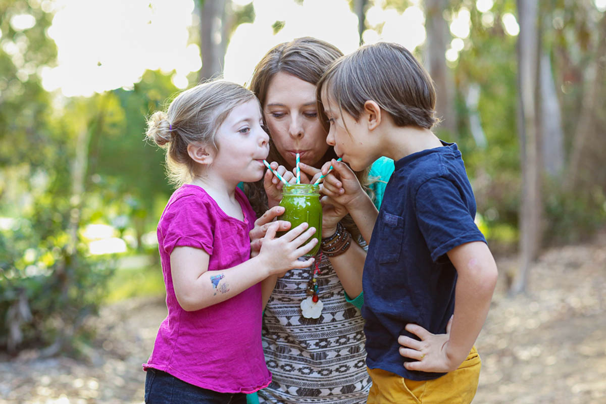 a mom and 2 kids all sipping a glass jar of green smoothie through blue striped paper straws.