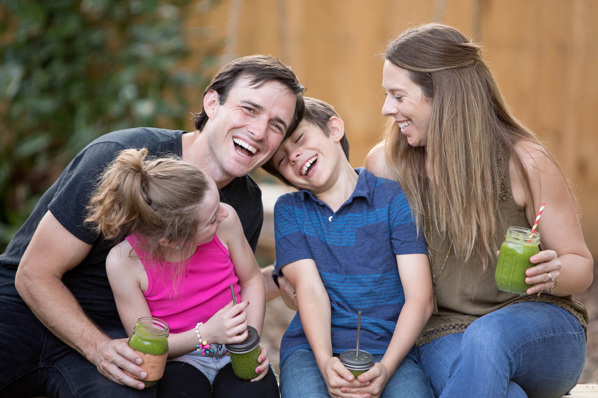 a family of four sitting and laughing together, each holding their own glass jar of vibrant green smoothie.