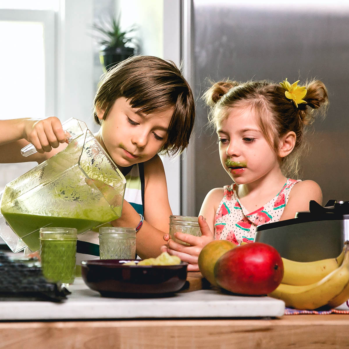 2 kids, one with a green smoothie mustache, pouring a blender pitcher of green smoothie into small jars on a counter full of fresh produce.