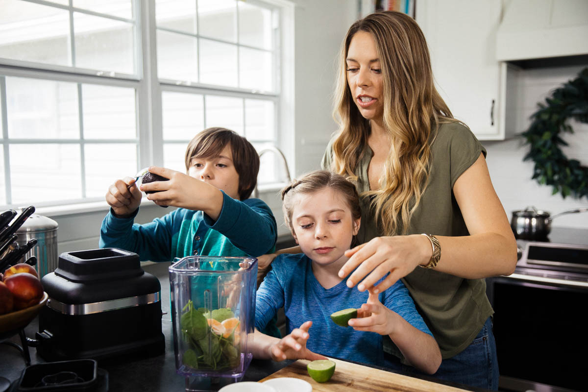 a mom and 2 kids in a kitchen putting avocados and limes into a blender pitcher.