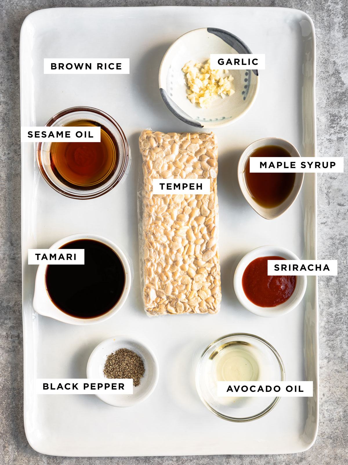 labeled ingredients for a tempeh bowl including brown rice, garlic, sesame oil, maple syrup, tempeh, tamari, sriracha, black pepper and avocado oil.