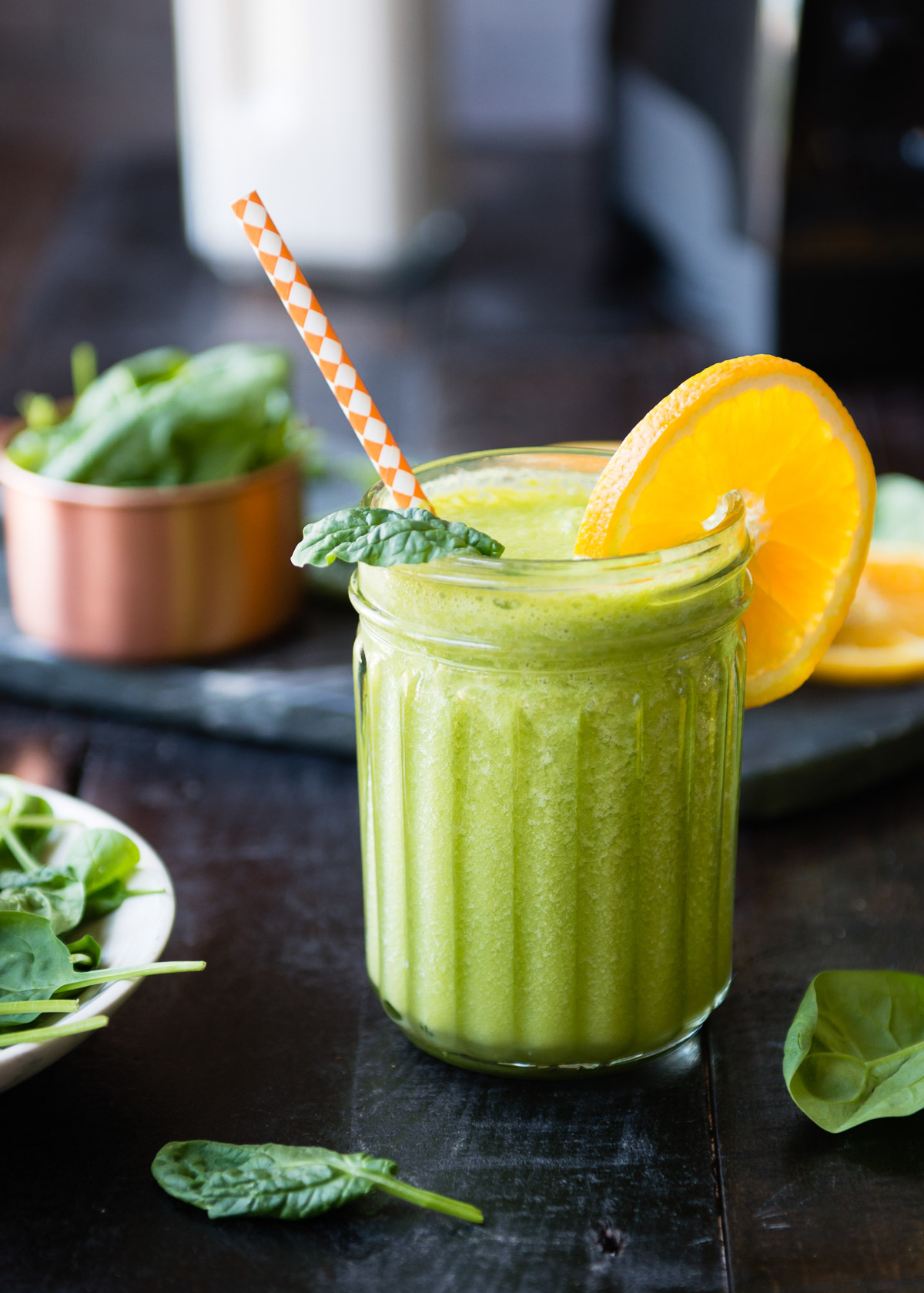 A fun textured Mason jar filled with spinach banana smoothie garnished with an orange slice and a paper straw.
