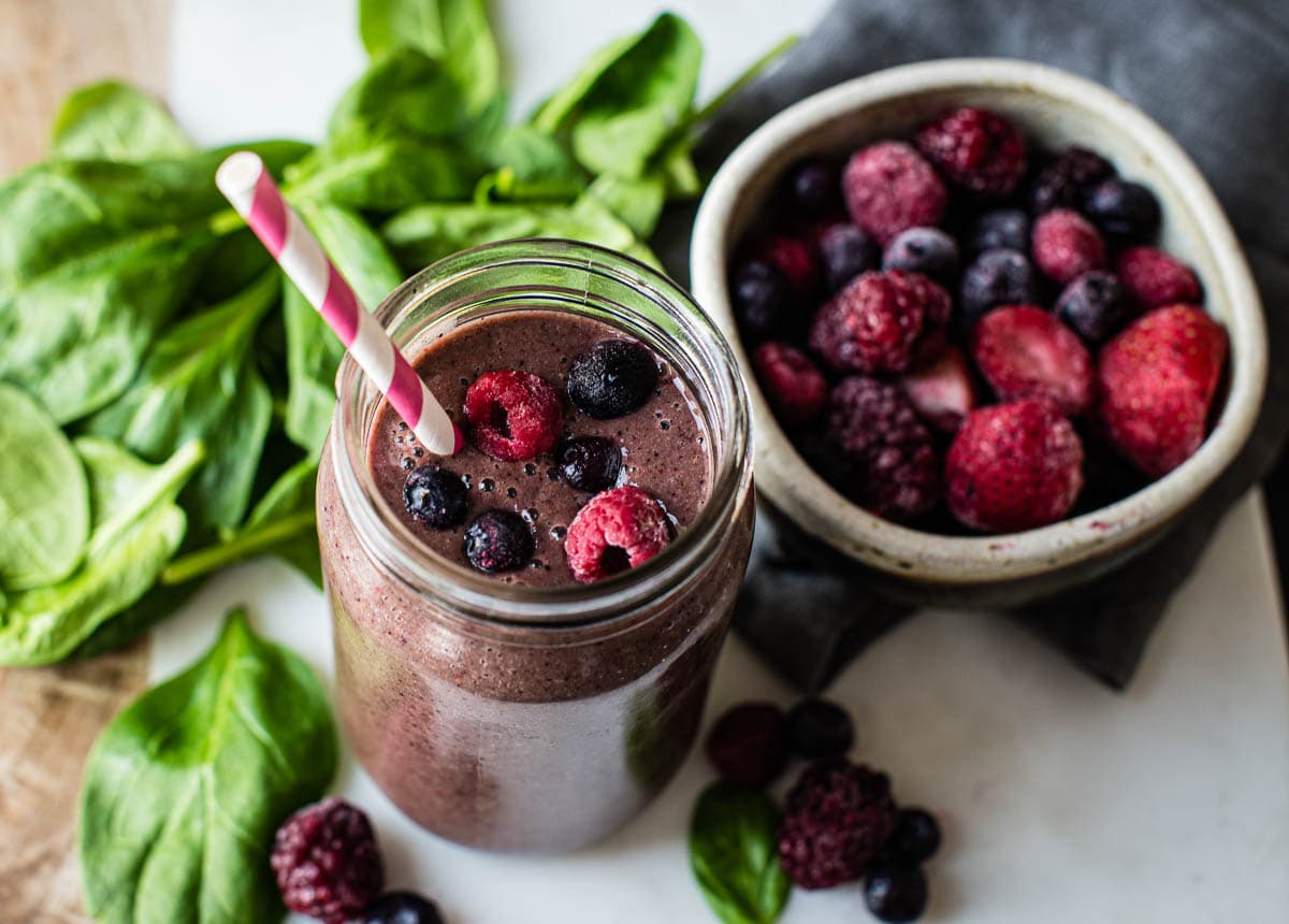 Healthy spinach berry smoothie in a glass jar, surrounded by whole ingredients like berries in a bowl and spinach on the counter top.