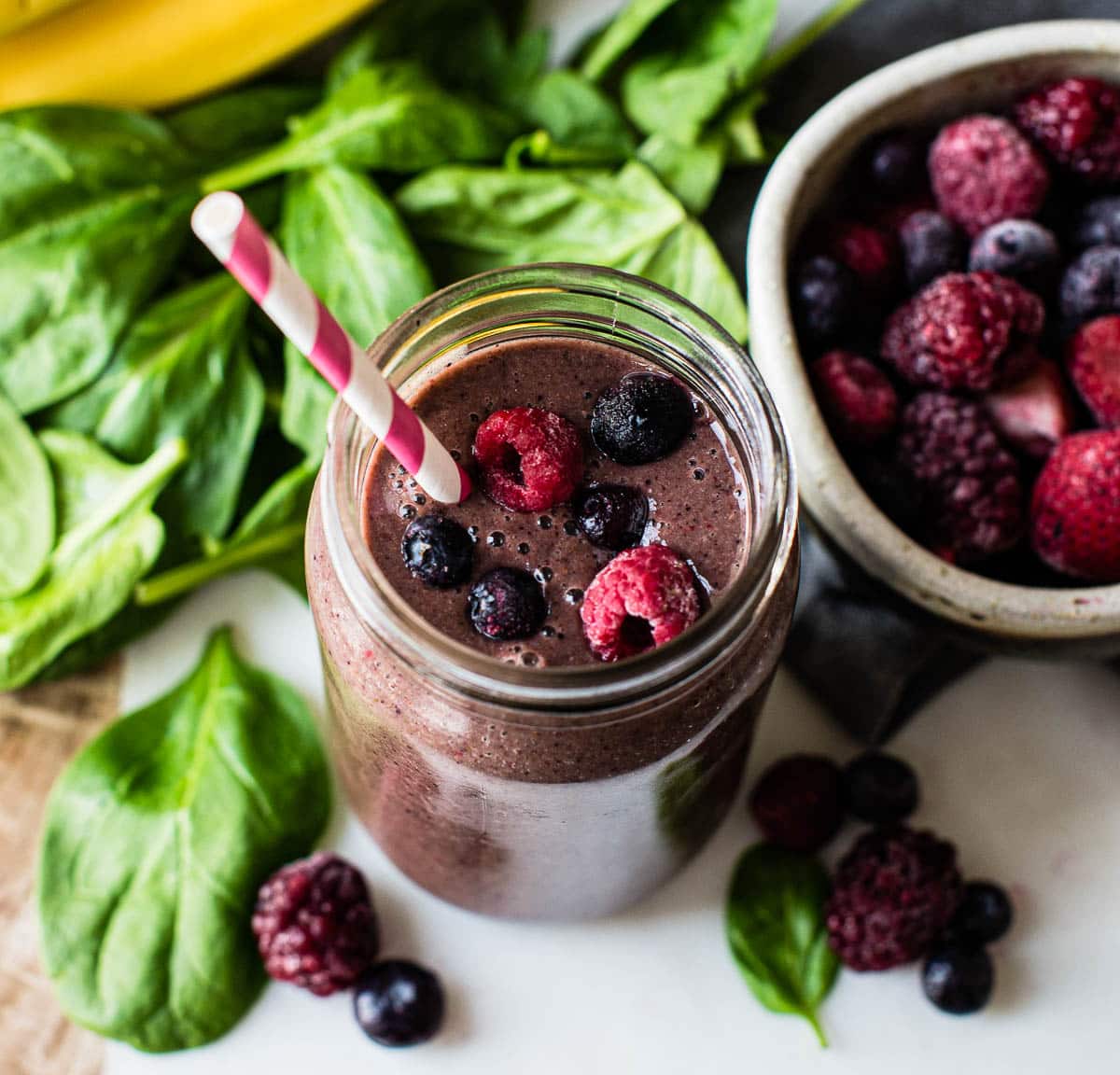 photo of spinach and berry smoothie in a glass jar with a paper straw. Frozen berries and fresh spinach are on the counter top surrounding the smoothie