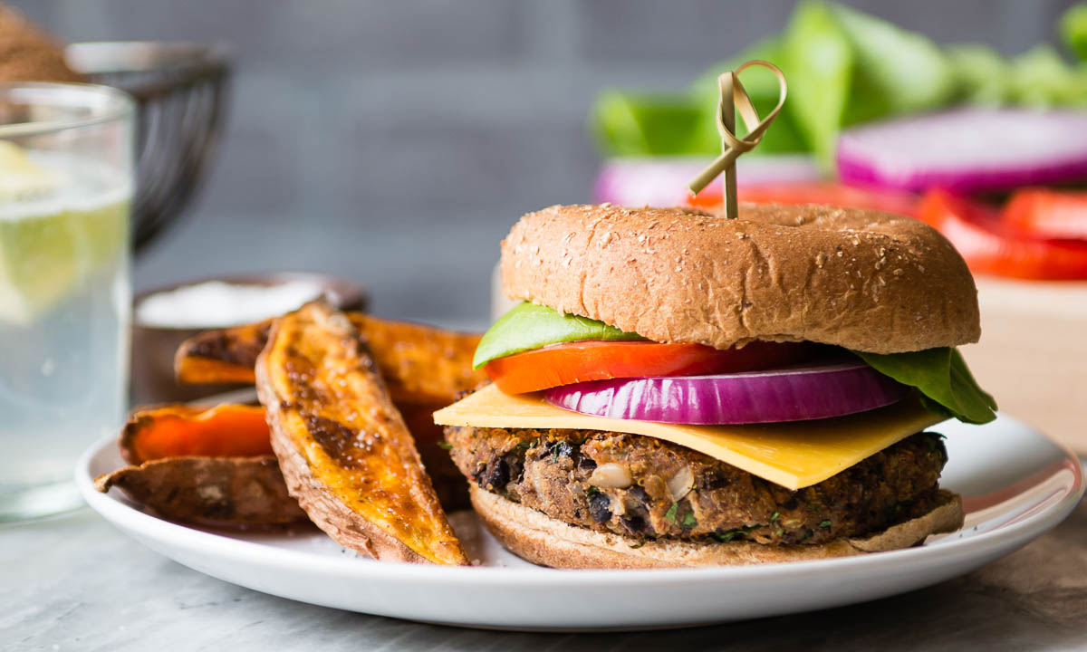 loaded black bean burger on a white plate with sweet potato wedges, a spring vegetarian recipe.