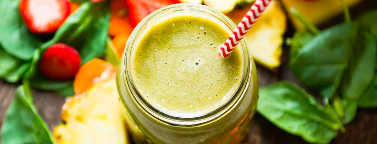green smoothie from above with a red and white straw; strawberries, pineapple, carrots, and spinach in the background