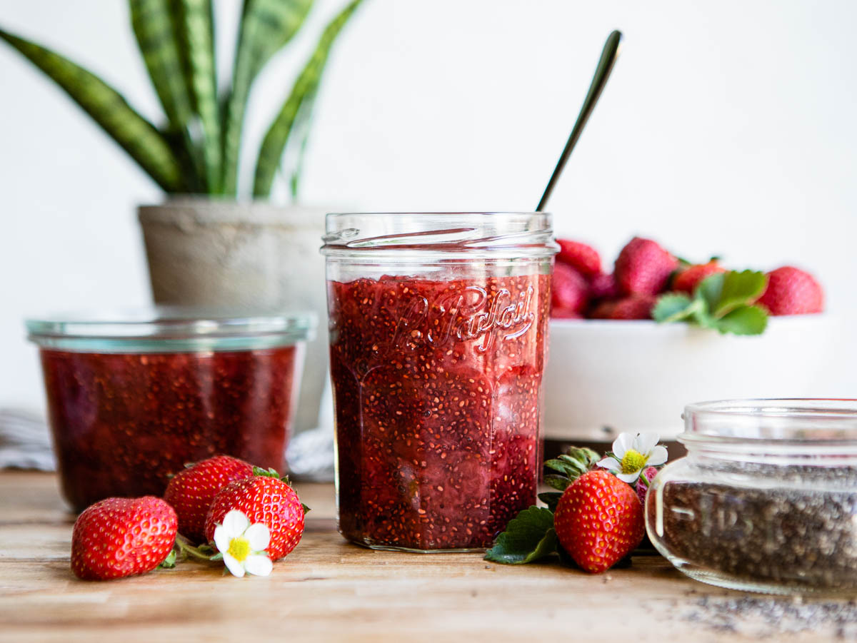 chia jam in 2 glass jars on a counter surrounded by fresh strawberries.