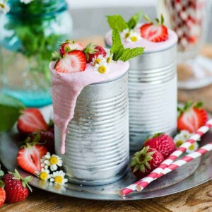 A refreshing and colorful display of two vegan strawberry milkshakes, garnished with fresh fruit and mint, with whole strawberries and straws arranged nearby.