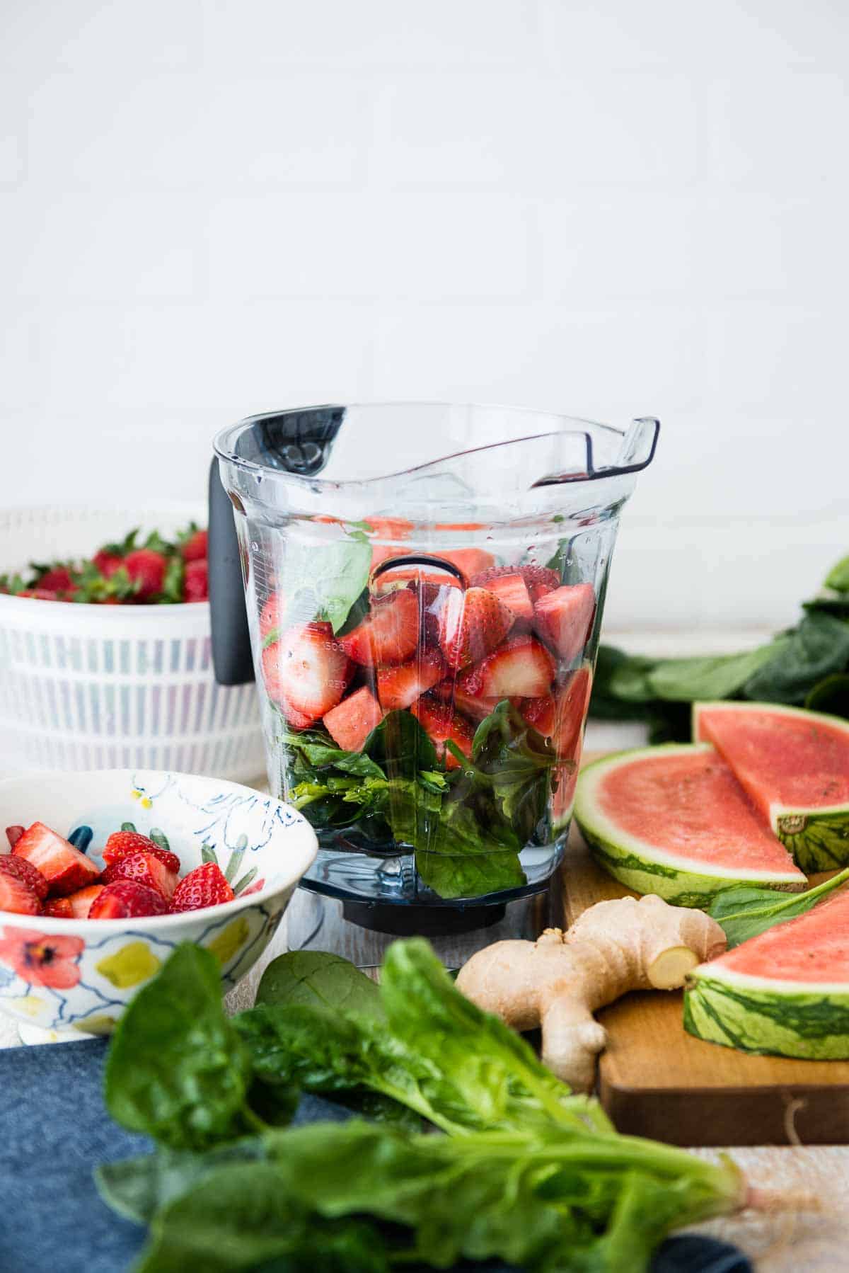 blender full of strawberries, spinach and water surrounded by other fresh fruits.