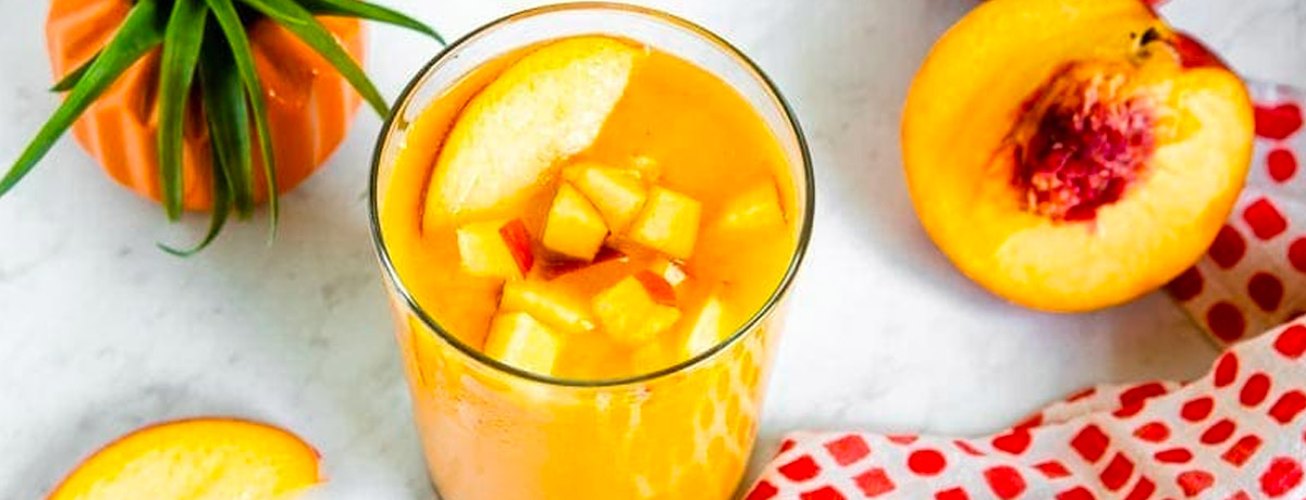 peach colored smoothie with fresh pieces of peach on top and a checkered cloth underneath