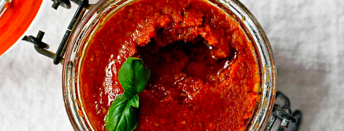 close up view of sun-dried tomato pesto in a jar, it looks deep red and has a sprig of basil on top.