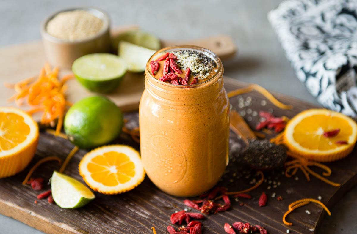 orange smoothie in a glass mason jar topped with goji berries, hemp hearts and chia seeds, on a wooden cutting board surrounded by citrus fruit.