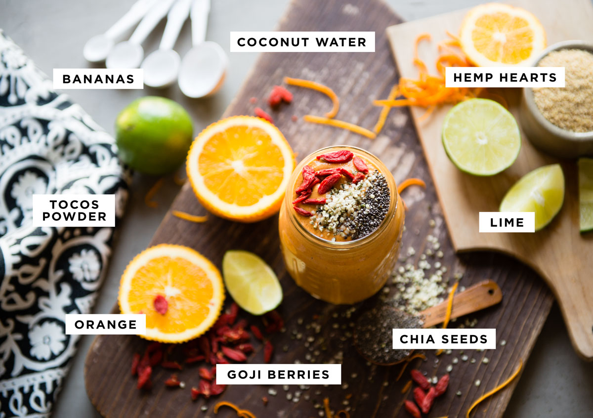 labeled ingredients for a superfood smoothie including coconut water, bananas, hemp hearts, lime, chia seeds, orange and tocos powder.