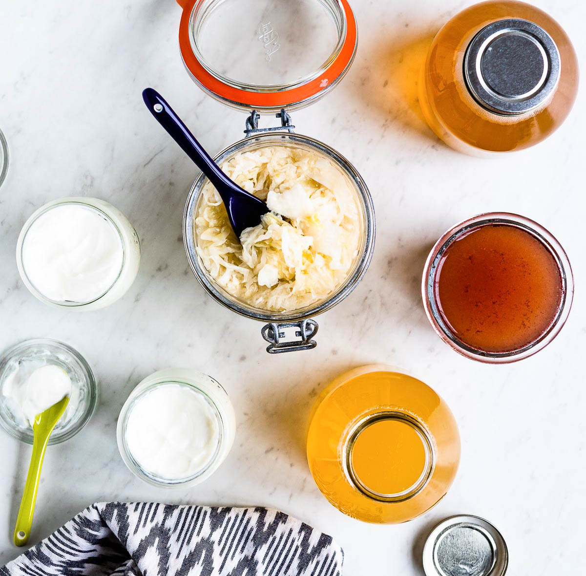 glass jar of sauerkraut surrounded by glass containers of kombucha and yogurt, all fermented superfoods.