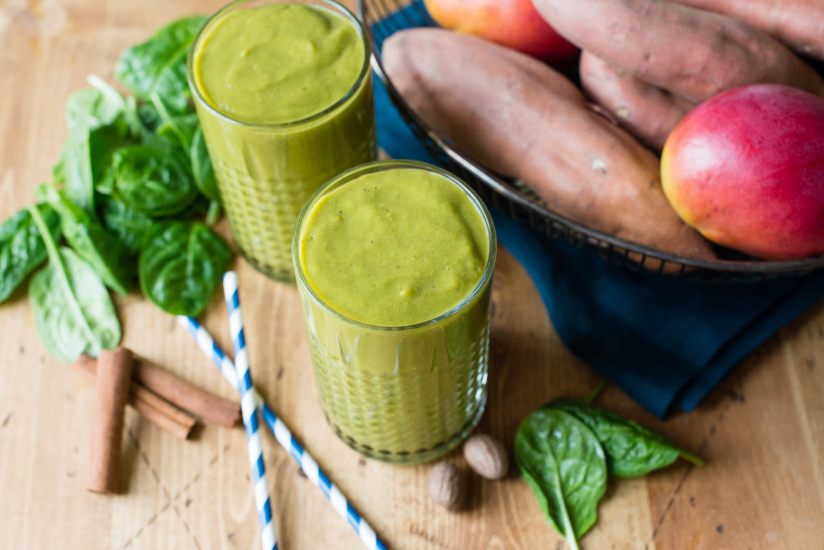 2 green smoothies in glasses on a wooden table with fresh spinach, cinnamon sticks, 2 striped blue straws and a basket of produce.