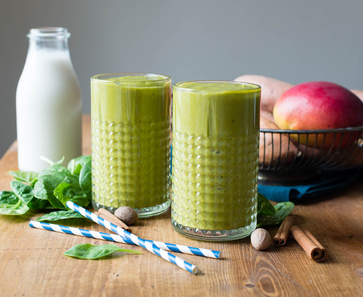 Two glasses of green smoothie sit on a wooden table, with blue striped straws. Fresh ingredients, including leafy greens and fruits, are placed nearby, adding healthy feel of the scene. 