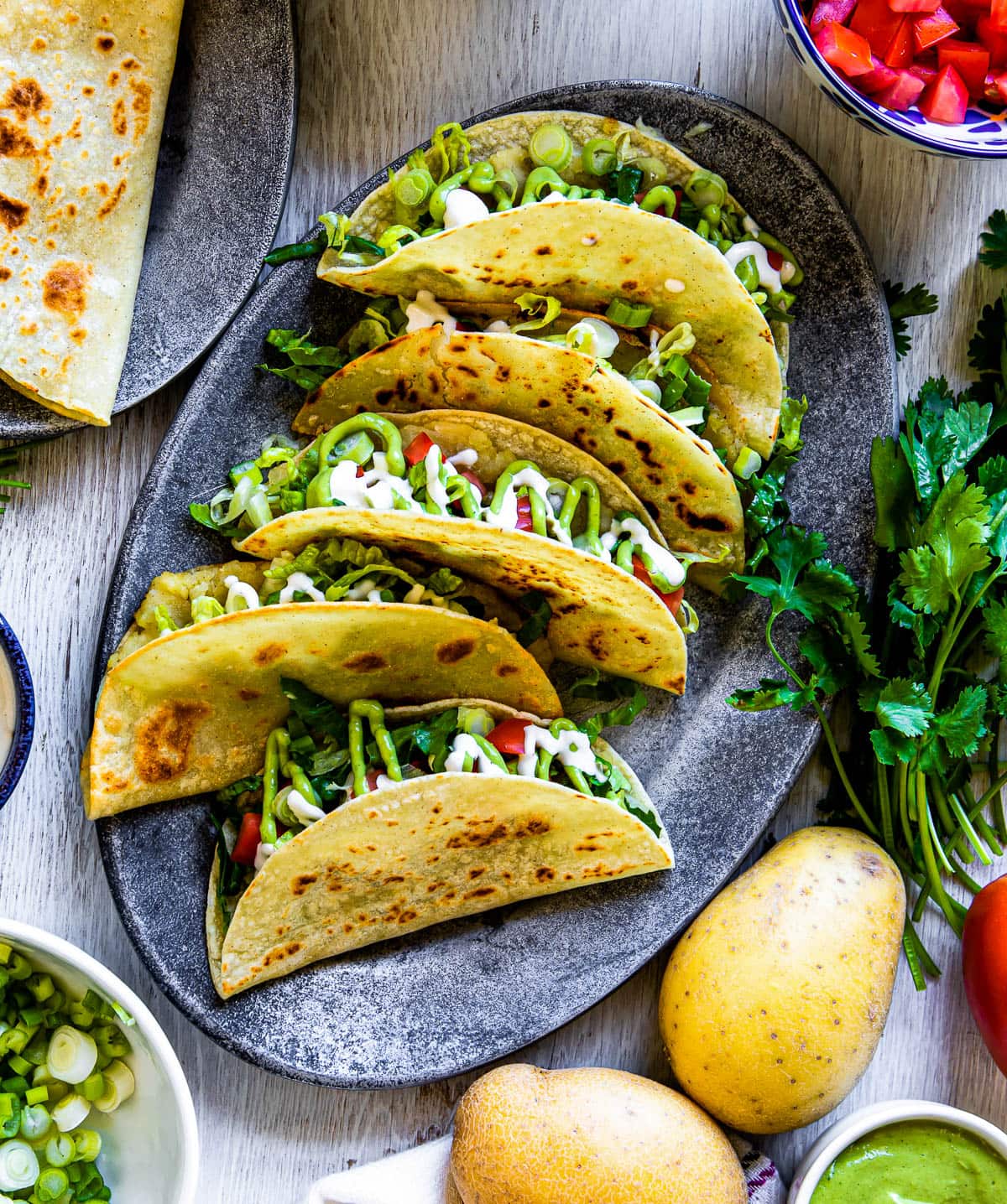 plate of corn tortilla tacos filled with potatoes and fresh ingredients on an oval plate, surrounded by cilantro and yellow potatoes.