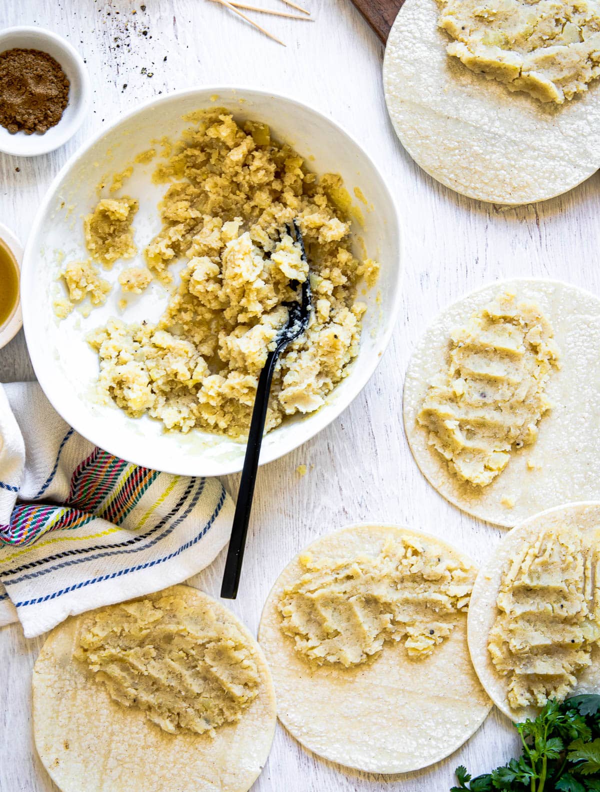 a white bowl of yellow mashed potatoes being added to 4 corn tortilla shells on a white surface.