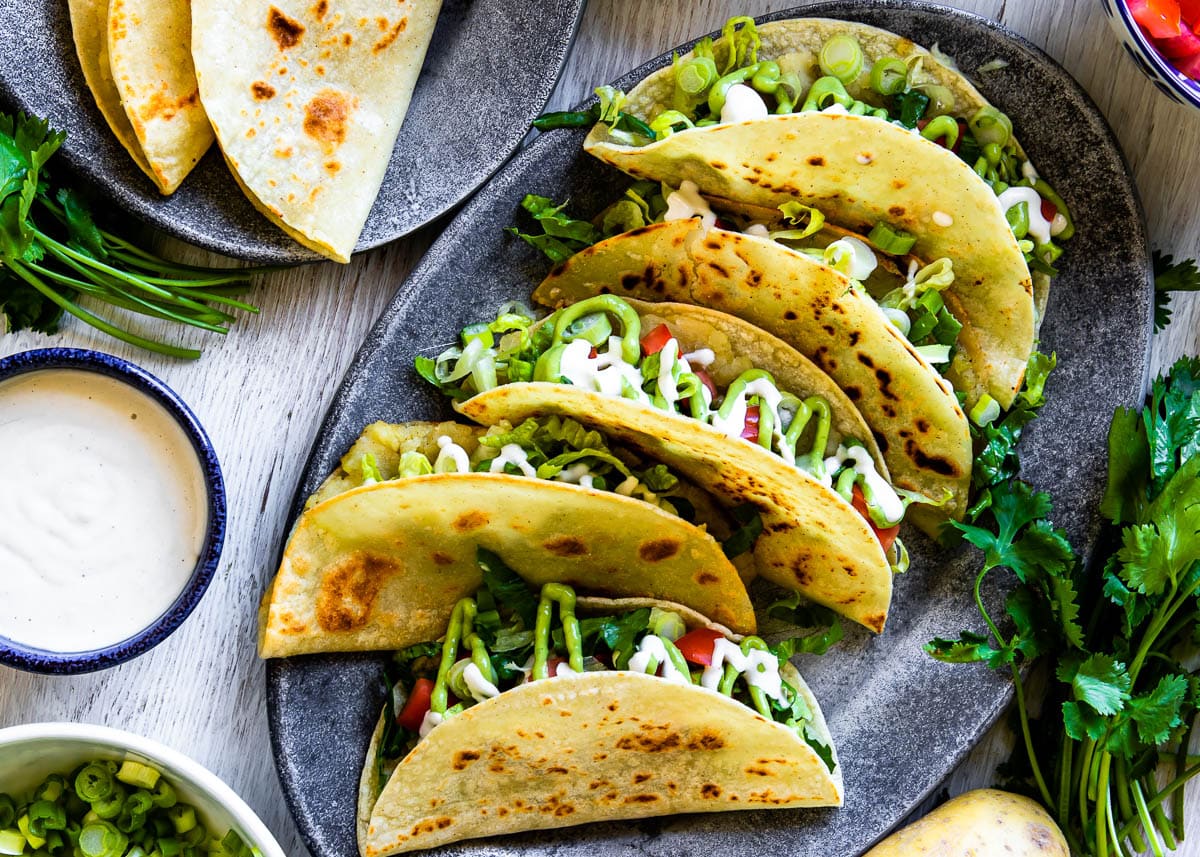 a gray, oval plate full of a Mexican-inspired street food, tacos de papa, topped with green avocado crema and white cashew cream.