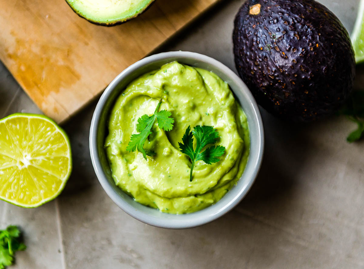 Bowl of avocado crema topped with fresh cilantro leaves and surrounded by avocado and lime ingredients. A perfect topping for crispy potato tacos.