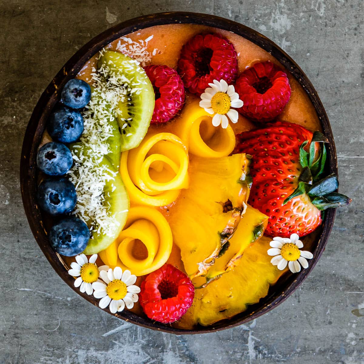 coconut bowl full of smoothie and topped with fresh blueberries, coconut flakes, kiwi, mango flowers, raspberries, pineapple, strawberries and white flowers.