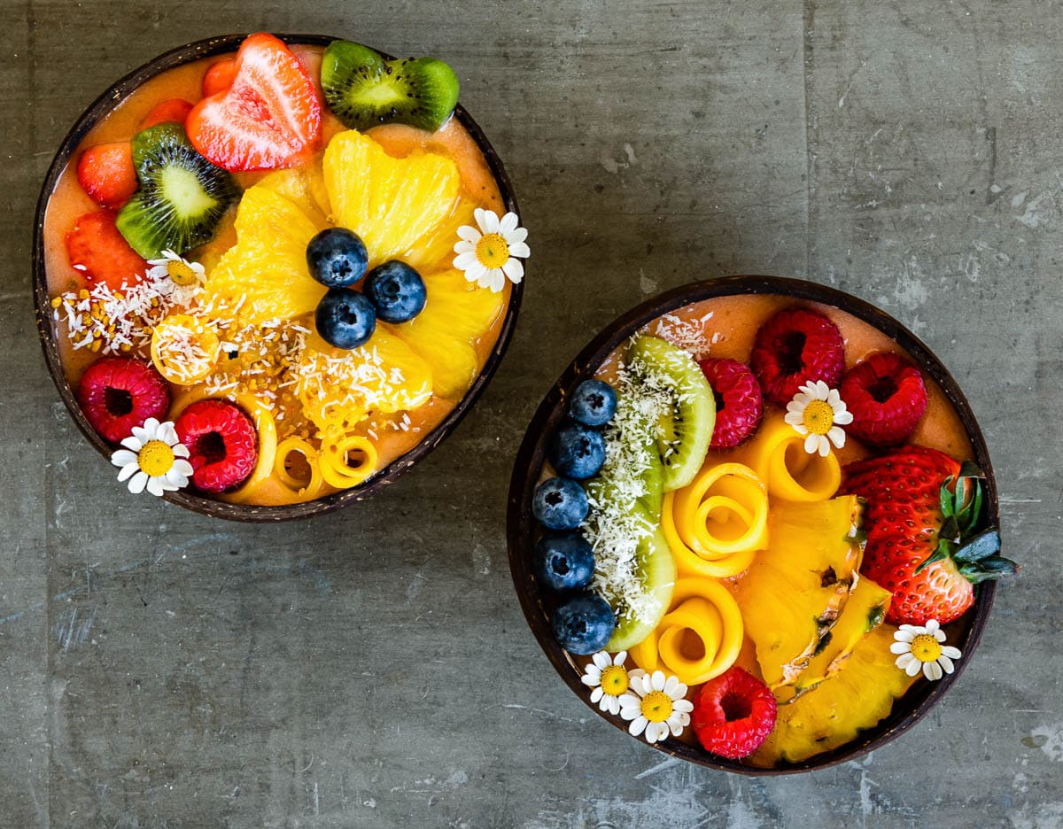overhead photo of 2 tropical smoothie bowls decorated with a variety of fresh fruit and some flowers