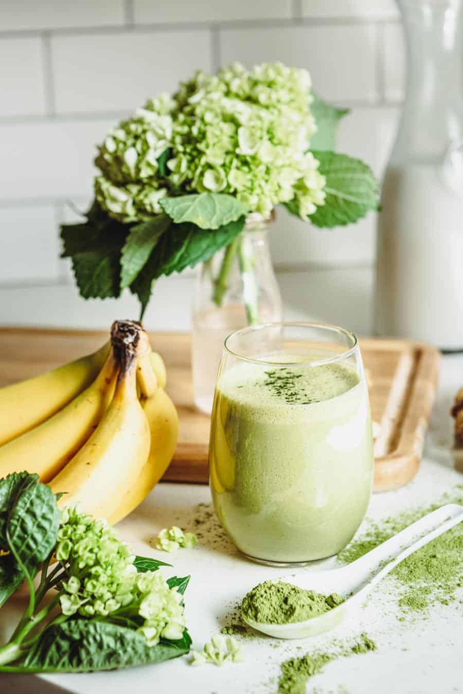 A matcha smoothie accompanied  by bananas, matcha powder and hydrangeas.