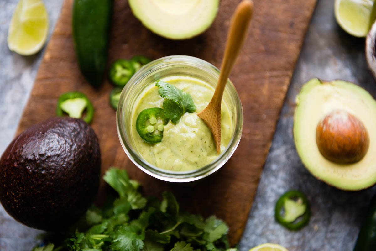 vegan avocado crema in a glass jar topped with cilantro, sliced jalapeño and a wooden spoon.